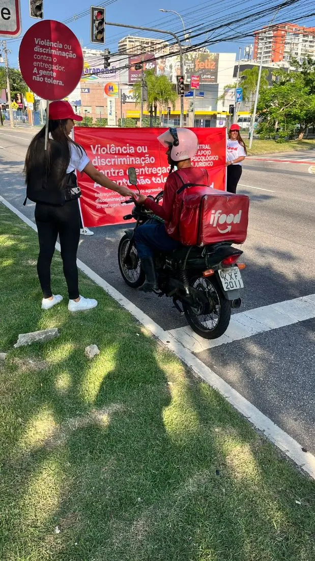 Representante de marca da Exfera Mídia com faixa iFood em sinal de trânsito entregando material a entregador